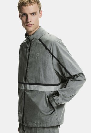 Fiatal férfi világosszürke szélálló dzsekiben, fekete szegélyekkel, egy kézzel a zsebében áll, egyszínű háttér előtt.