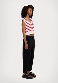 Top de punto sin mangas y recortado con rayas horizontales rojas sobre un fondo crema, combinado con pantalones negros holgados y sandalias negras gruesas.