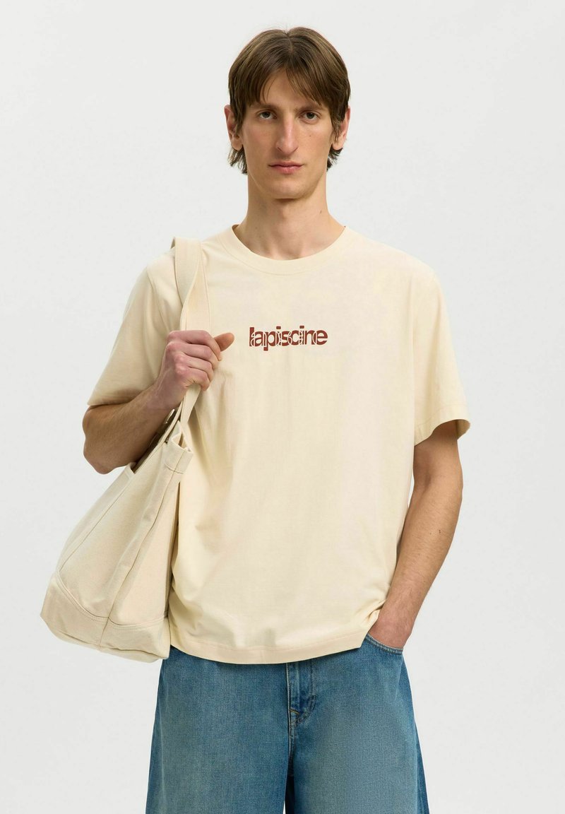 Jeune homme aux cheveux bruns debout portant un t-shirt beige avec le texte "lapiscine", un jean bleu et un grand sac beige en bandoulière.