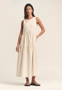 Ljus beige, ärmlös maxiklänning med lagerdesign och samlade detaljer. Slät textur, enkel rund halsringning och ankellång nederkant.