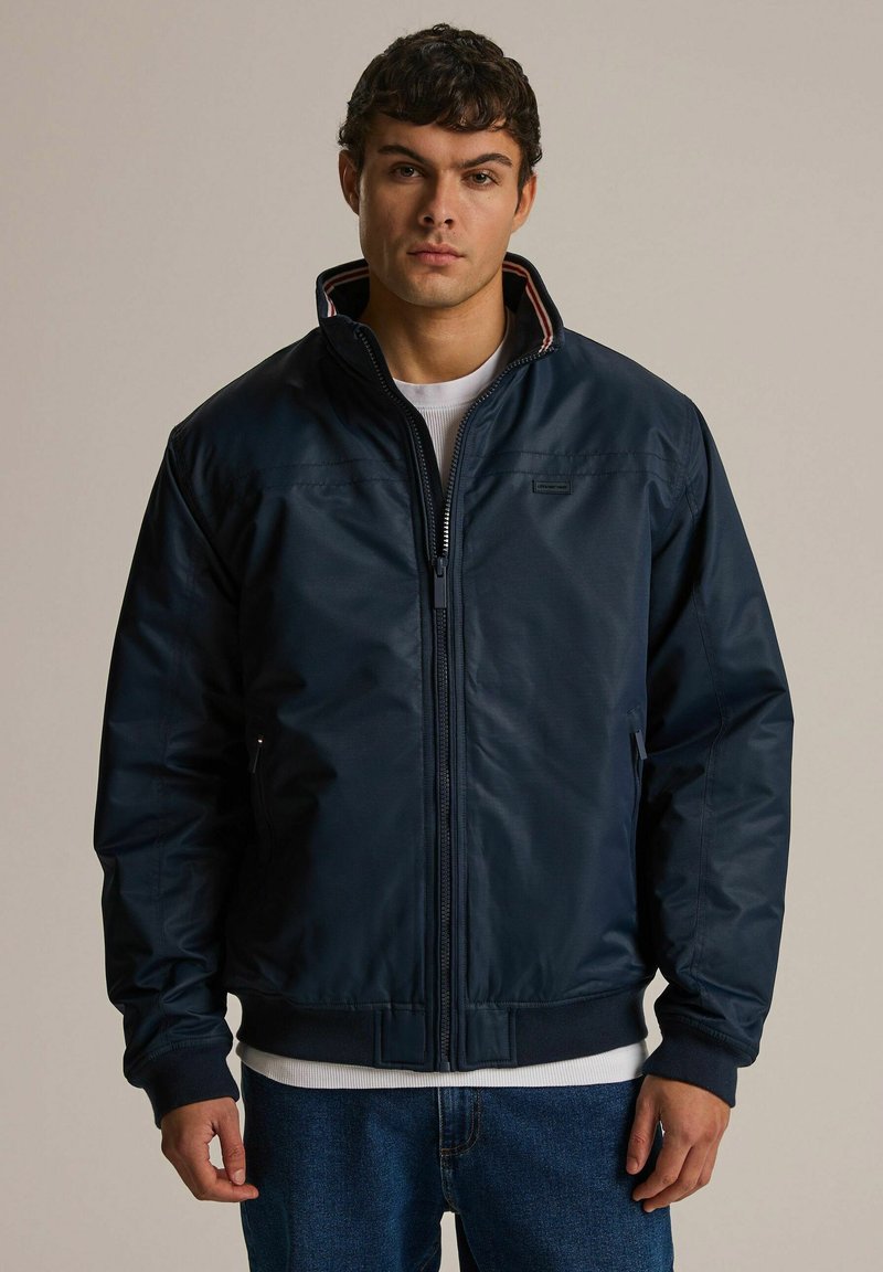 Granatowa kurtka ocieplana z wysokim kołnierzem, zamkiem błyskawicznym z przodu i prążkowanymi mankietami. Posiada subtelne logo na piersi oraz paskowane podszycie.