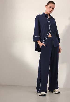 Femme debout, une main dans la poche, portant une chemise ample bleu marine boutonnée et un pantalon large assorti avec un liseré blanc, baskets blanches.