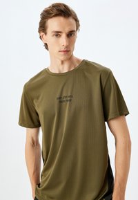 Camiseta de manga corta en verde oliva, hecha de un tejido ligero y transpirable con un cuello redondo, que presenta una frase motivacional impresa en negro.