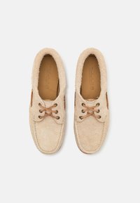 Zapatos de ante beige con una textura suave, puntera redondeada, acentos en cuero marrón y cordones marrones, con un diseño clásico de zapato náutico.