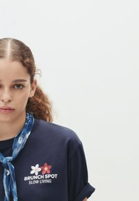 Mørkeblå t-shirt med oplagte ærmer; har broderet tekst "BRUNCH STED SLOW LIVING" og to blomster i rød og hvid. Blå tørklæde som tilbehør.