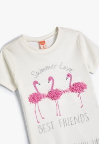 Camiseta de manga corta color crema con tres flamencos rosas con acentos texturizados, y el texto "Amor de Verano Mejores Amigos" en plata.