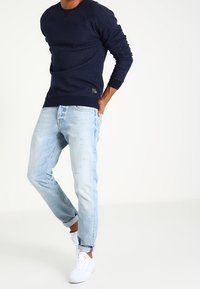 Sudadera azul marino de mangas largas y puños acanalados, combinada con vaqueros ajustados azul claro y zapatillas blancas, de pie contra un fondo blanco.