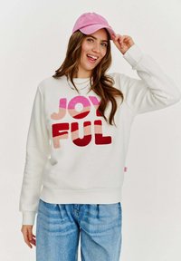 Biele mikina s veľkými, textúrovanými písmenami "JOYFUL" v odtieňoch ružovej a červenej. Skombinovaná so svetlomodrými džínami a ružovou čiapkou.