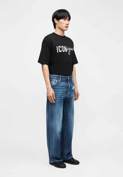 Άνδρας που στέκεται, φοράει μαύρο μπλουζάκι με την επιγραφή "ICONique", φαρδιά μπλε τζιν με ελαφριά φθορά και μαύρα παπούτσια, μπροστά σε απλό ανοιχτόχρωμο φόντο.