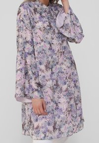 Tunique à motif floral avec des manches longues évasées. Couleurs comprenant le violet, le rose et le gris. Dotée d'un col rond et de détails de boutons.