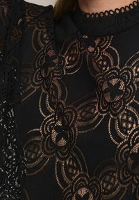 Schwarzes Spitzentop mit aufwendigen Blumenmustern, einem strukturierten Design und einem hohen Kragen mit gewellten Rändern.