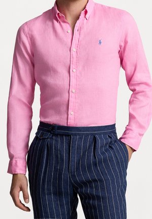 Mężczyzna noszący jaskraworóżową koszulę na guziki, zestawioną z granatowymi, wysokim stanem spodniami w drobną prążkę z bocznymi guzikami do regulacji.