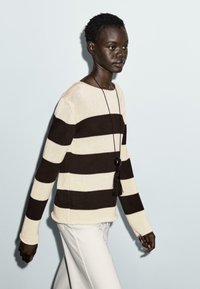 Knit sweater with beige and dark brown horizontal stripes, round neckline, and long sleeves, paired with white trousers and a pendant necklace.