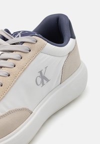 Witte sneaker met beige suede accenten, grijs logo, grijze veters en een marineblauwe gewatteerde kraag. Glad materiaal en een dikke witte zool.