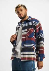 Kostkovaná flanelová košile v námořnické modré, červené a bílé barvě s knoflíkovým zapínáním, náprsní kapsou a strukturovaným materiálem; kombinována se šedým tričkem.