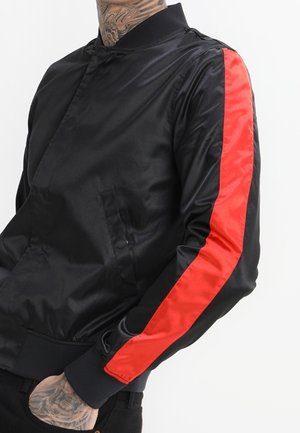 Veste en satin noire avec une bande rouge audacieuse le long de la manche portée par une personne tatouée avec la main dans la poche.