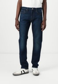 Jeans blu scuro con vestibilità dritta, design a cinque tasche, cuciture a contrasto e chiusura con bottone metallico. Indossati con sneakers bianche.