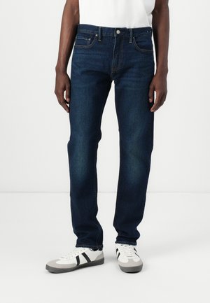 Jeans oscuros de corte recto, diseño de cinco bolsillos, costuras en contraste y cierre de botón metálico. Llevados con zapatillas blancas.