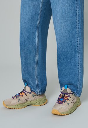 Sneakers beige con lacci multicolori, suole verde maculate e dettagli blu, abbinati a jeans blu larghi.