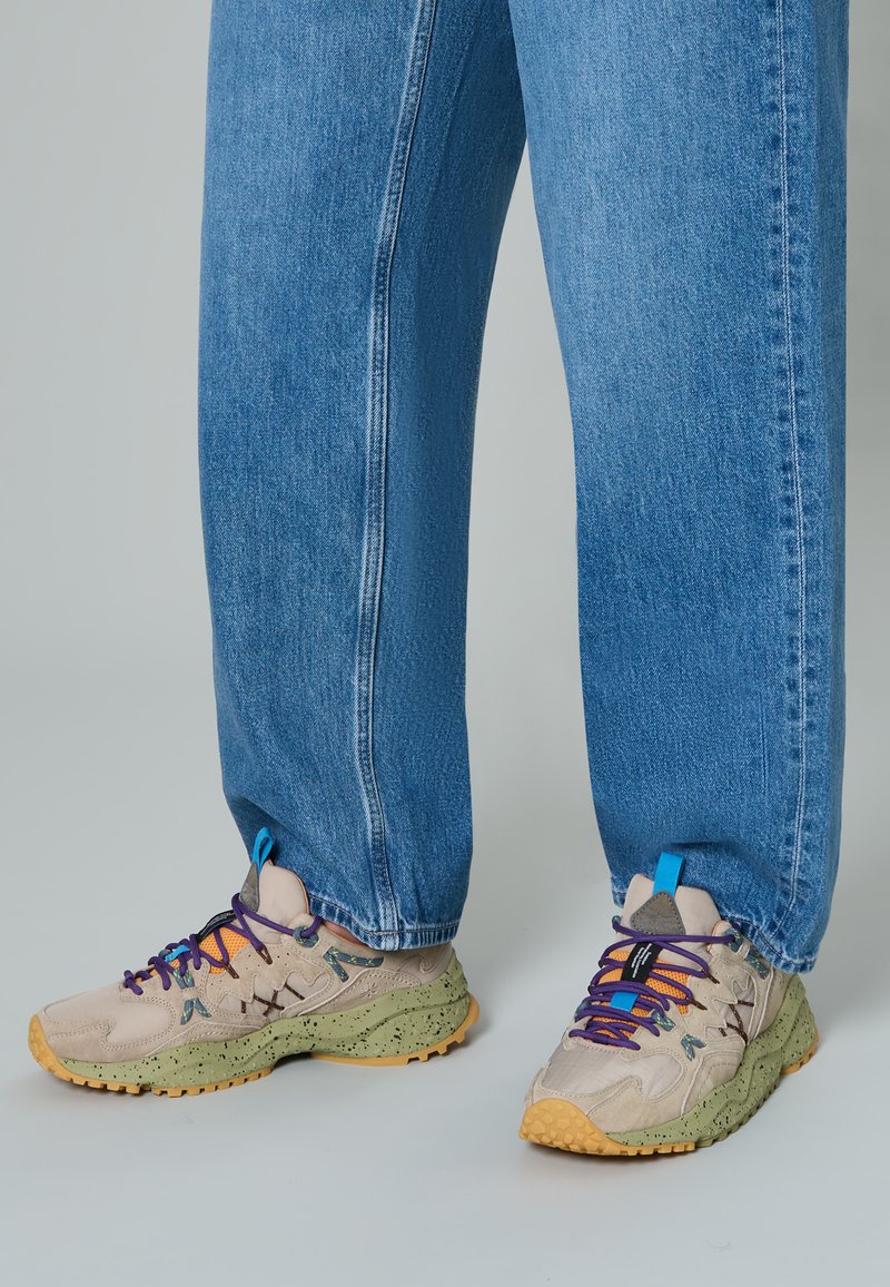Sneakers beige con lacci multicolori, suole verde maculate e dettagli blu, abbinati a jeans blu larghi.