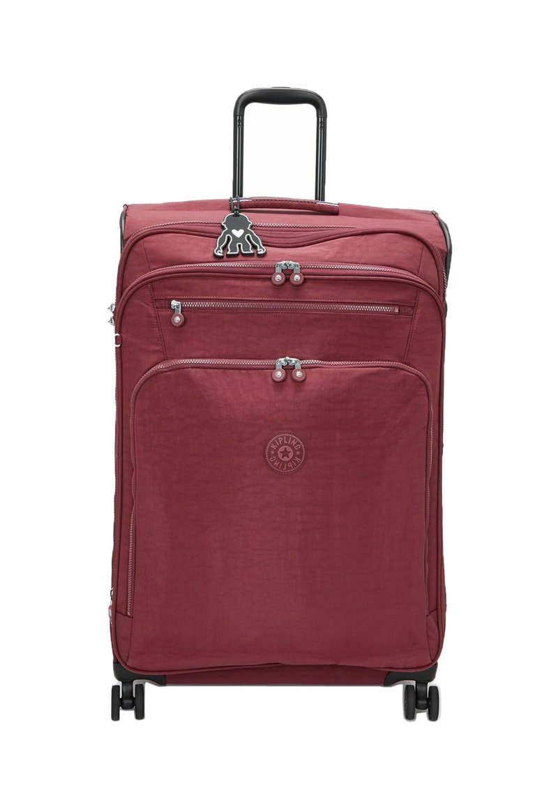 Red textile suitcase with a retractable handle, two front zippered pockets, and four wheels. Features a logo and a decorative charm.
