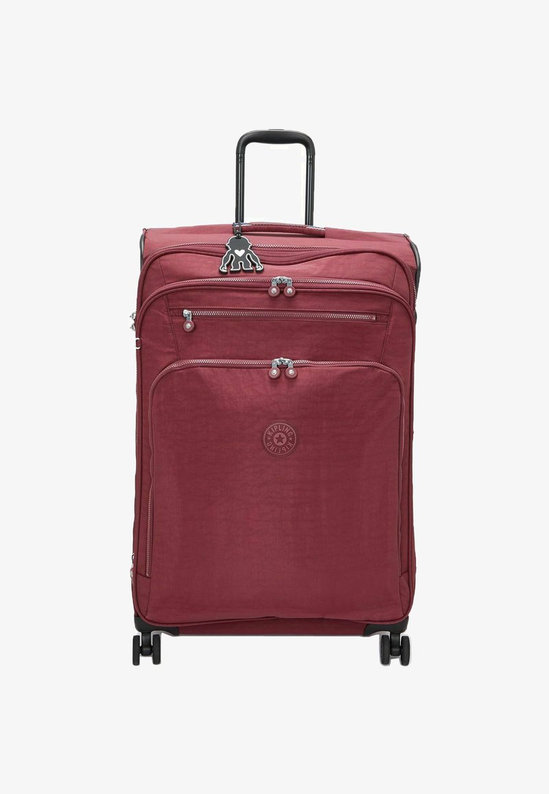 Red textile suitcase with a retractable handle, two front zippered pockets, and four wheels. Features a logo and a decorative charm.