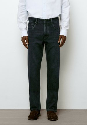 Uomo con jeans scuri a gamba dritta, camicia bianca a maniche lunghe infilata e mocassini in camoscio marrone, in piedi contro un muro semplice.