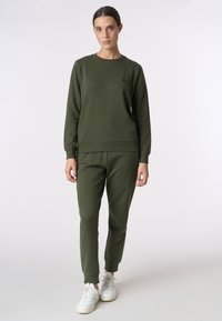 Felpa verde oliva con maniche lunghe e polsini a coste, abbinata a pantaloni tuta affusolati dello stesso colore. Sneakers bianche completano il look.