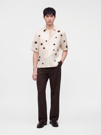 Jeune homme portant une chemise crème à manches courtes avec des motifs floraux marron, un pantalon marron foncé et des mocassins foncés, debout avec une main dans la poche.