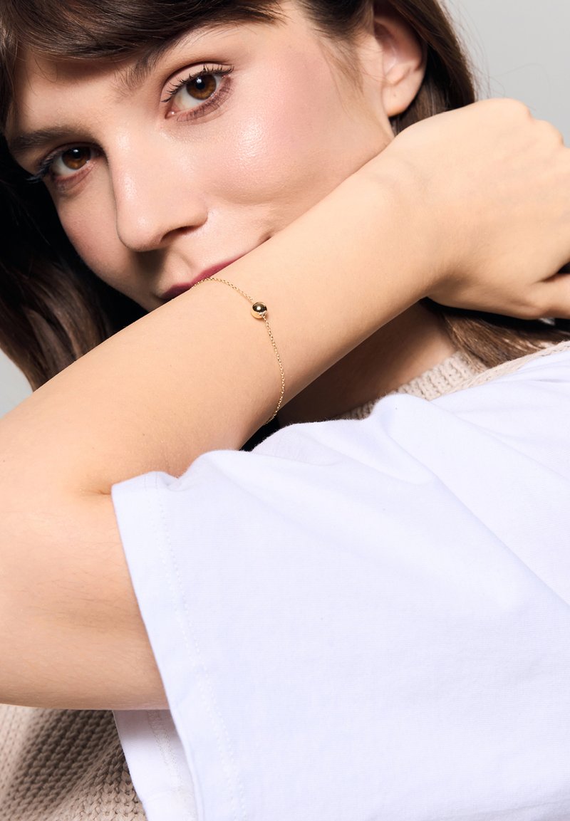 Złoty bransoletka z małą okrągłą kulką, z delikatnym łańcuszkiem. Noszona na nagim nadgarstku na jasnym tle.