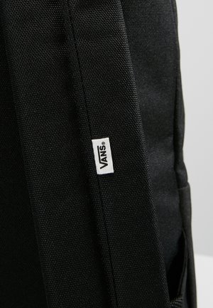 Close-up van een zwarte Vans-rugzakriem met een klein wit Vans-logo label dat in de stof is genaaid.