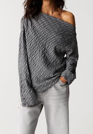 Kvinde iført grå ternet off-shoulder langærmet top og lysegrå revnede jeans, poserer med den ene hånd i lommen på ensfarvet baggrund.