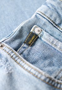 Jasnoniebieskie dżinsy z wyraźną metką Superdry, żółtym szwem i srebrnym guzikiem przy przedniej kieszeni.