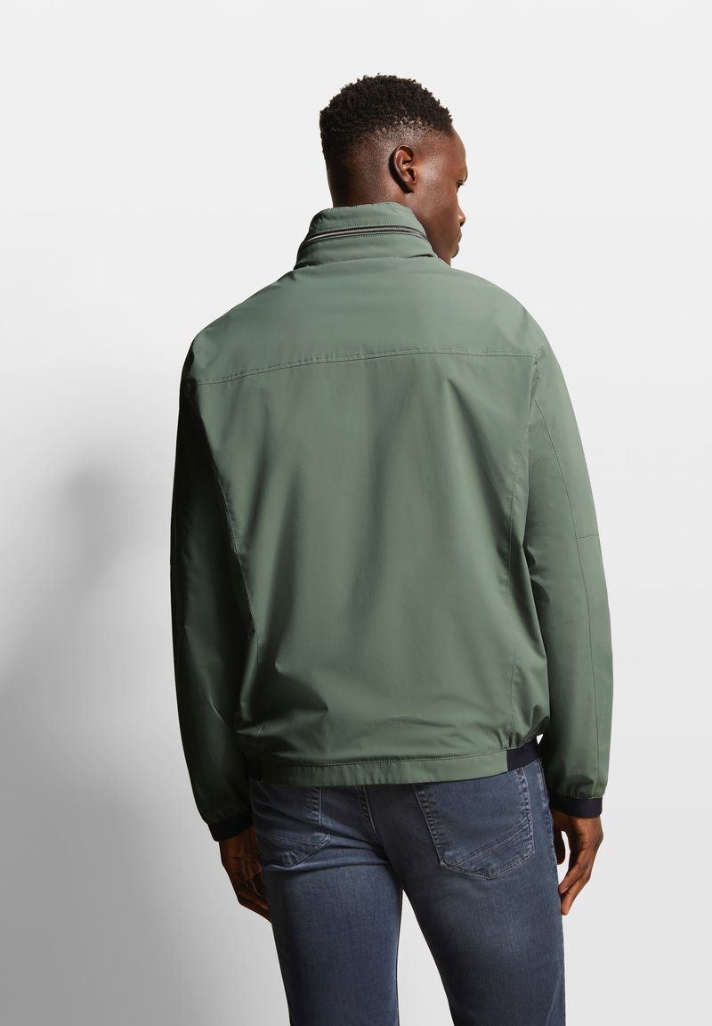 Veste légère verte avec un col haut, texture lisse, poignets côtelés et fermeture éclair, associée à un jean en denim.