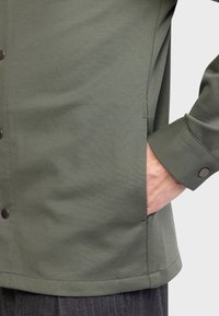 Camisa verde de botones, hecha de una tela suave, con un bolsillo lateral y puños con detalle de botón. Dobladillo recto.