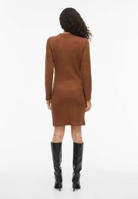 Abito marrone aderente al ginocchio con maniche lunghe e una trama sottile, abbinato a stivali neri alti in pelle con tacco.