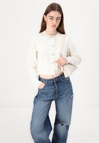 Cream cropped cardigan with textured pattern and large buttons, paired with high-waisted blue jeans featuring a ripped detail.