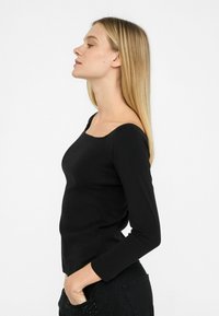 Schwarzes Langarmoberteil mit quadratischem Ausschnitt, aus softem Stoff gefertigt. Mit tailliertem Design und minimalistischer Markenpräsenz auf der Brust.