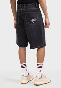 Sorte denim shorts med en afslappet pasform, der har baglommer og et broderet 'REBEL' grafik. Sammen med hvide sokker og beige sneakers.