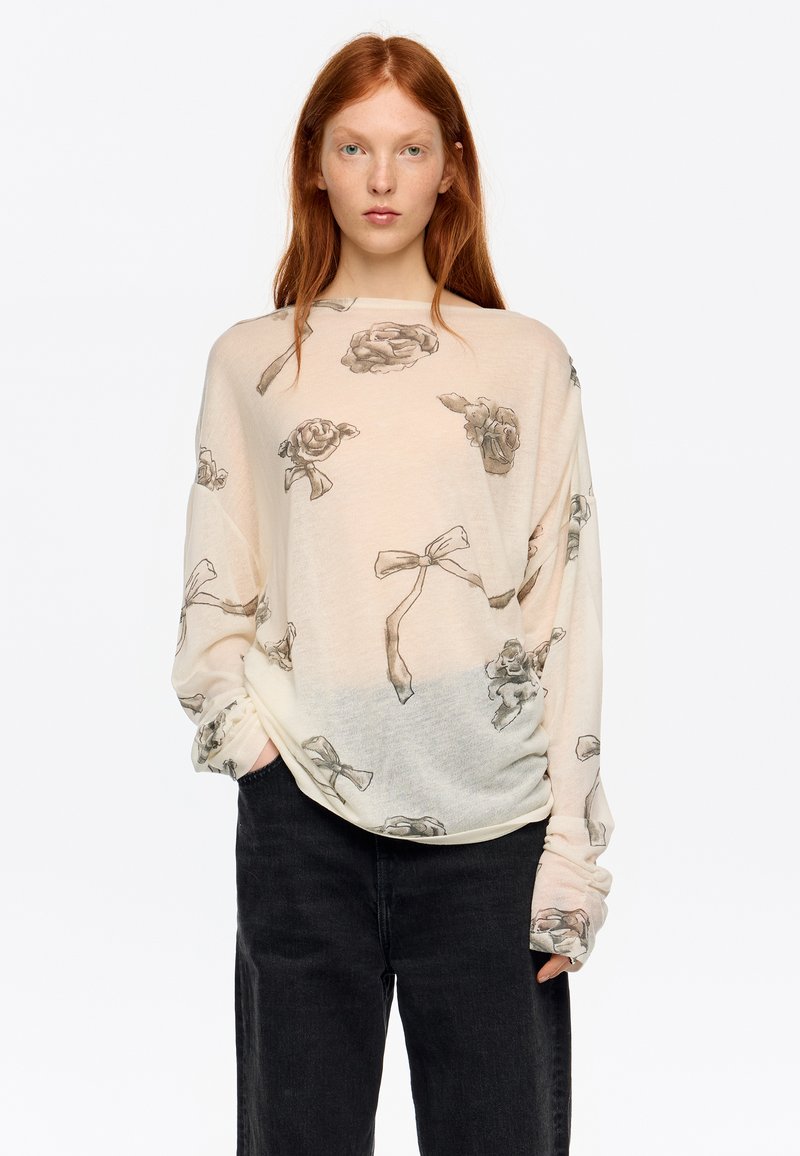 Jasnobeżowa koszula z długim rękawem, z nadrukiem kwiatów i kokardek, wykonana z miękkiego materiału, luźnego kroju, z podwiniętymi rękawami. W połączeniu z czarnymi spodniami.