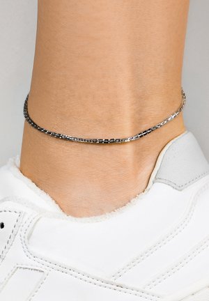 Chaîne de cheville en argent portée autour d'une cheville au-dessus d'une basket blanche avec des détails et des coutures grises.