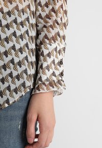 Blusa a maniche lunghe realizzata in tessuto leggero, con un motivo ripetuto di teste di cavallo in tonalità marrone scuro e beige, con polsini arricciati.