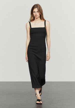 Giovane donna che cammina con un vestito midi nero aderente senza maniche e sandali neri su uno sfondo chiaro uniforme.