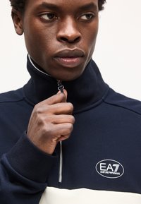 Άνδρας με μπλε ναυτικό και λευκό πλεκτό πάνω μέρος με φερμουάρ, τραβάει το φερμουάρ κοντά στο πηγούνι του, εμφανές το λογότυπο EA7 Emporio Armani στο στήθος.