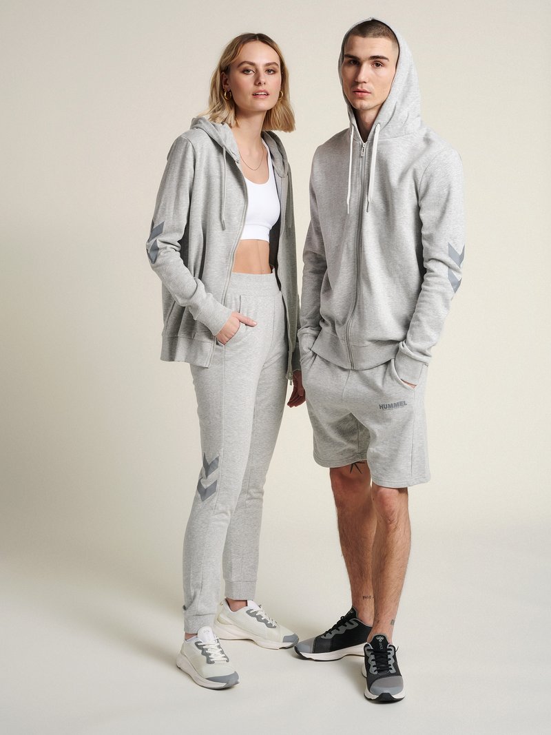Junge Frau und Mann tragen passende graue Kapuzenpullover, Jogginghosen und Sneakers, stehen nebeneinander vor neutralem Hintergrund.