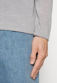 Grå långärmad skjorta i mjukt stickat tyg som är kombinerad med ljusblå denimjeans. Nærbild av en hand som vilar på skjortans underkant.