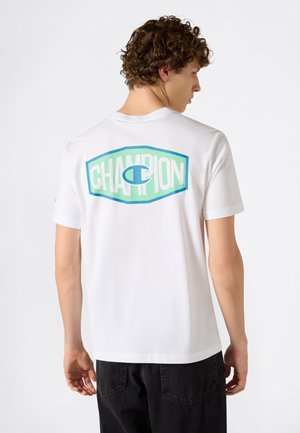 Giovane uomo con capelli ricci che indossa una maglietta bianca con il logo "CHAMPION" blu e verde sulla schiena, abbinata a pantaloni neri.