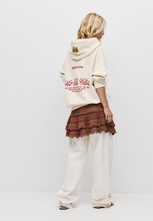 Felpa con cappuccio panna con testo rosso e etichetta verde, abbinata a una gonna all'uncinetto multicolore. Pantaloni bianchi ampi completano il look.