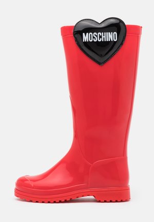 Høj blank rød regnstøvle med sort hjerte label nær toppen, der viser Moschino-mærket i hvidt.