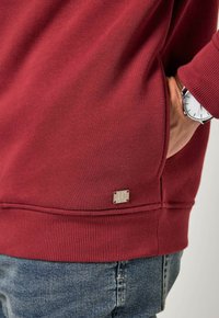 Sudadera burdeos de textura suave, con puños acanalados y una pequeña etiqueta de logo plateado en el dobladillo. Combinada con jeans azules y un reloj.